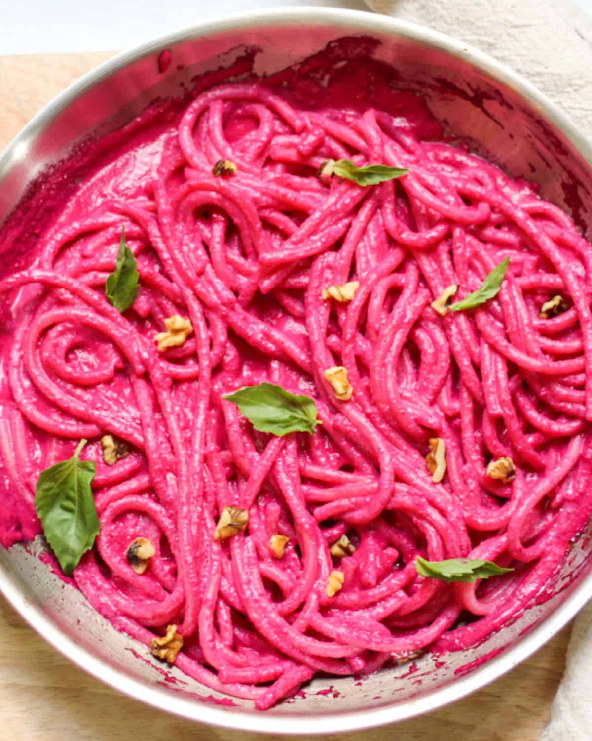 Spaghetti in a pink beetroot sauce in a skillet garnished with walnuts and fresh basil leaves.