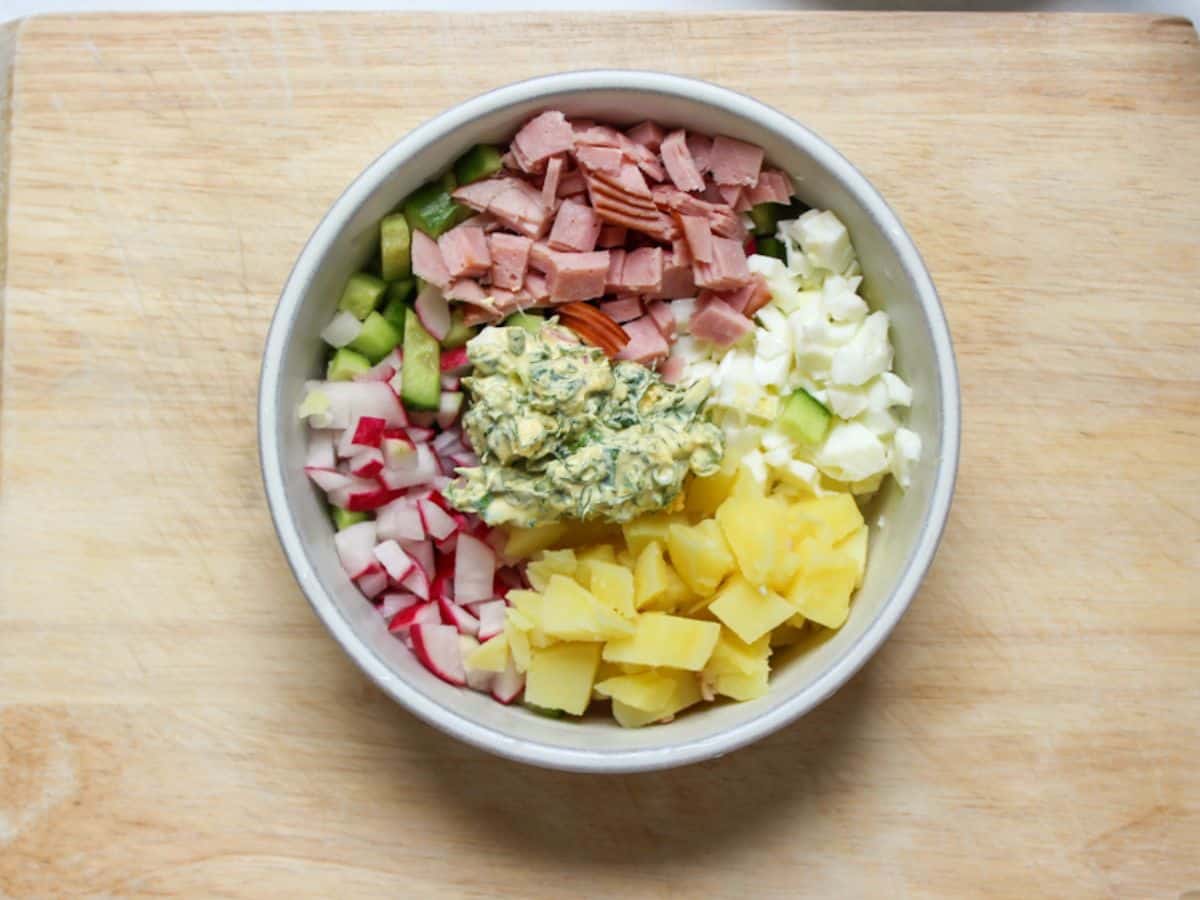 Diced potatoes, radishes. cucumbers, ham, egg whites and dill mixture in a white bowl.