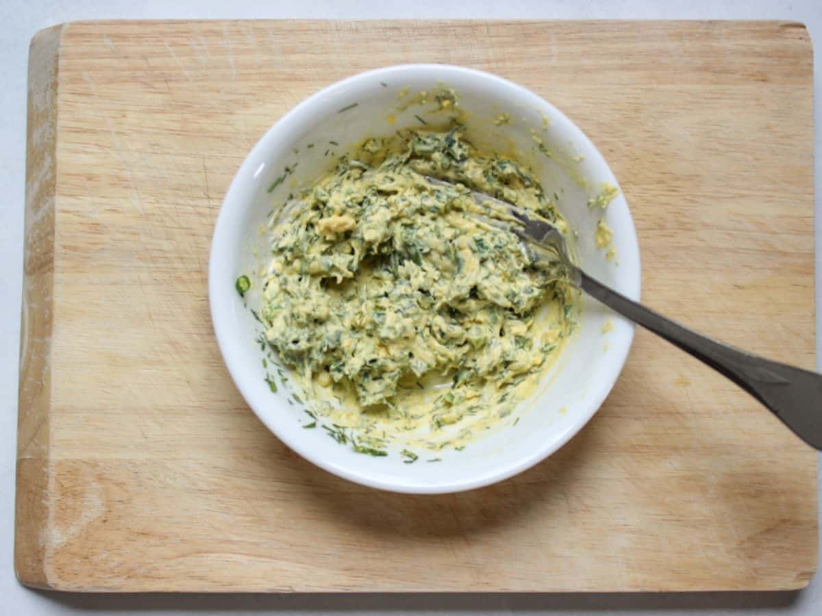 Mixture of egg yolks, mustard, sour cream, dill and green onions in a white small bowl with a fork in it.