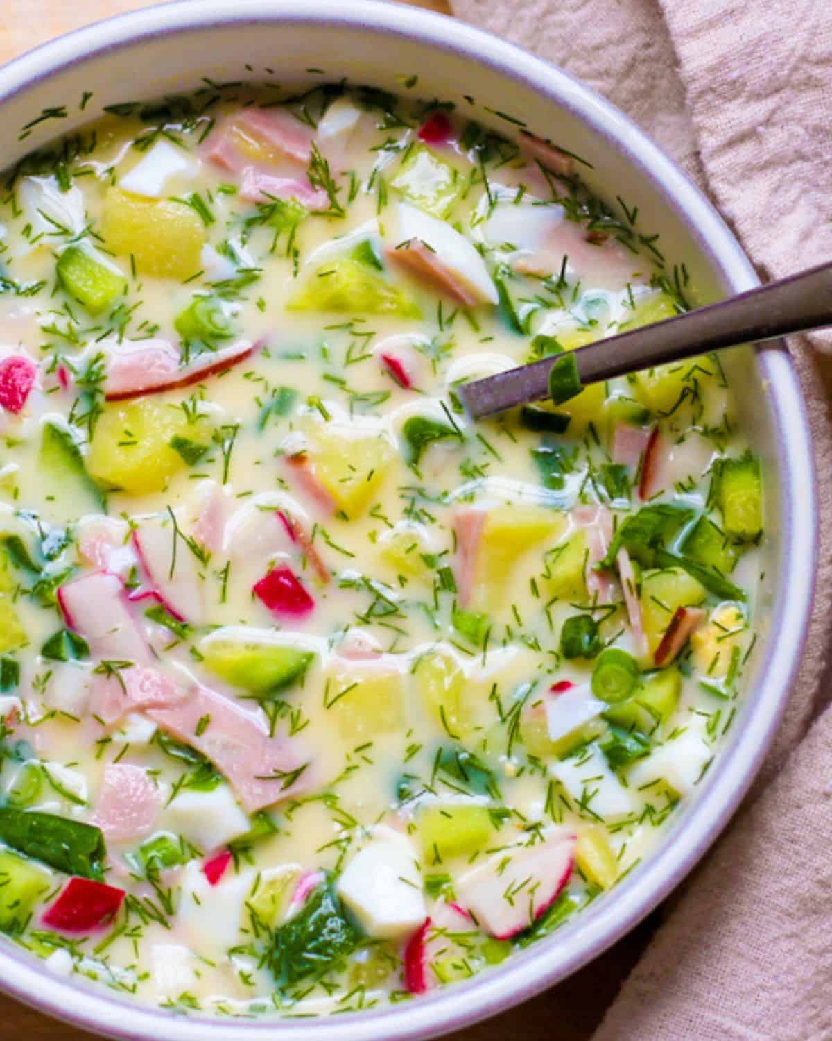 Okroshka cold soup with diced meat, radishes, cucumbers, dill in a deep white bowl.