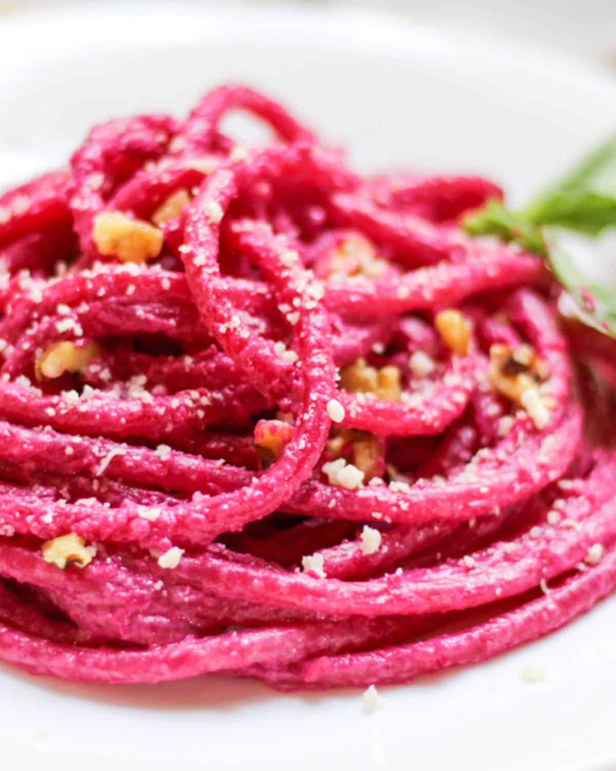 Beetroot spaghetti served on a white plate is topped with walnut pieces and grated Parmesan cheese. 