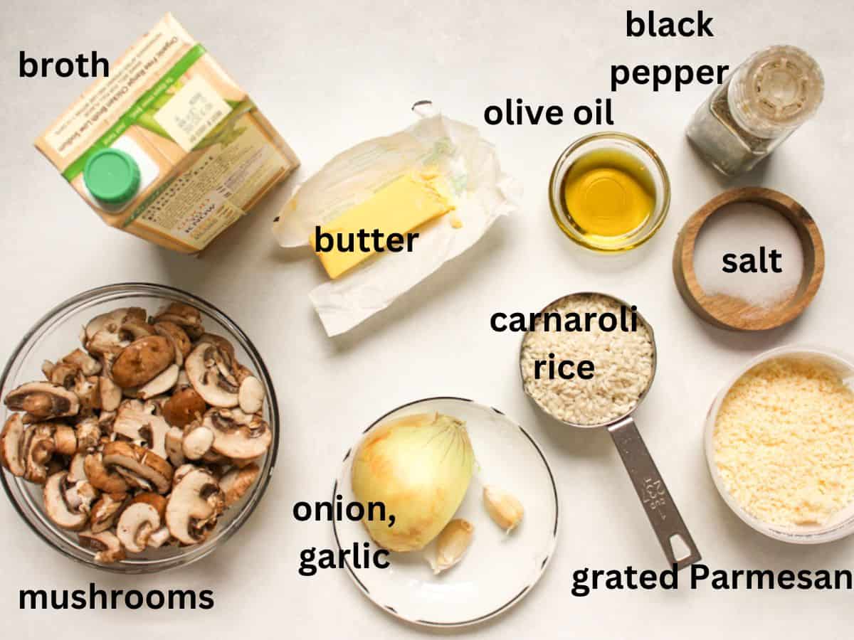 Recipe ingredients on a white surface. Broth, mushrooms, butter, onion, garlic, carnaroli rice, olive oil, salt, pepper, grated cheese.