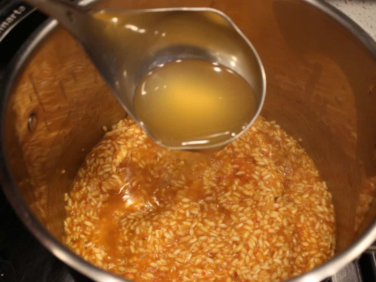 A ladle with broth poured into the pot with cooking risotto.