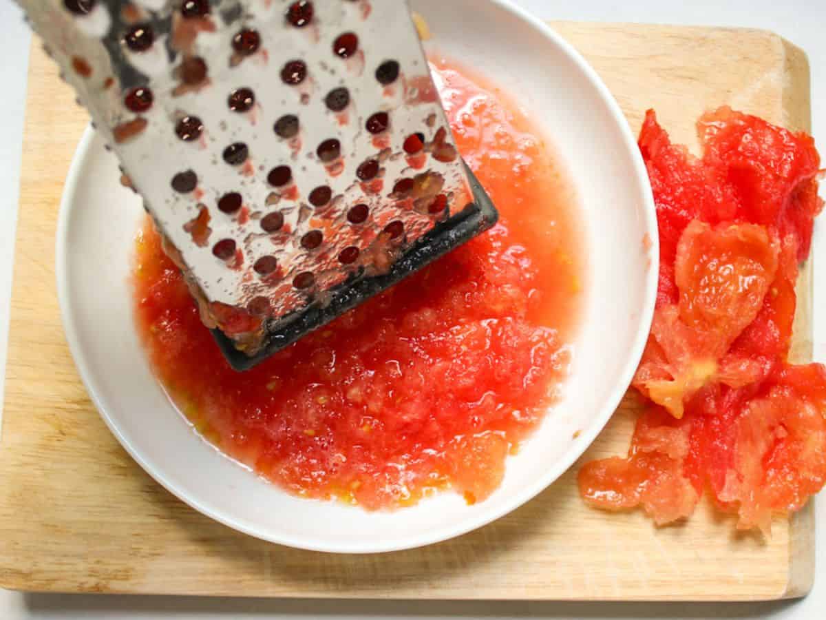Fresh tomatoes grated into a puree with stainless steel box grater in a white dish.