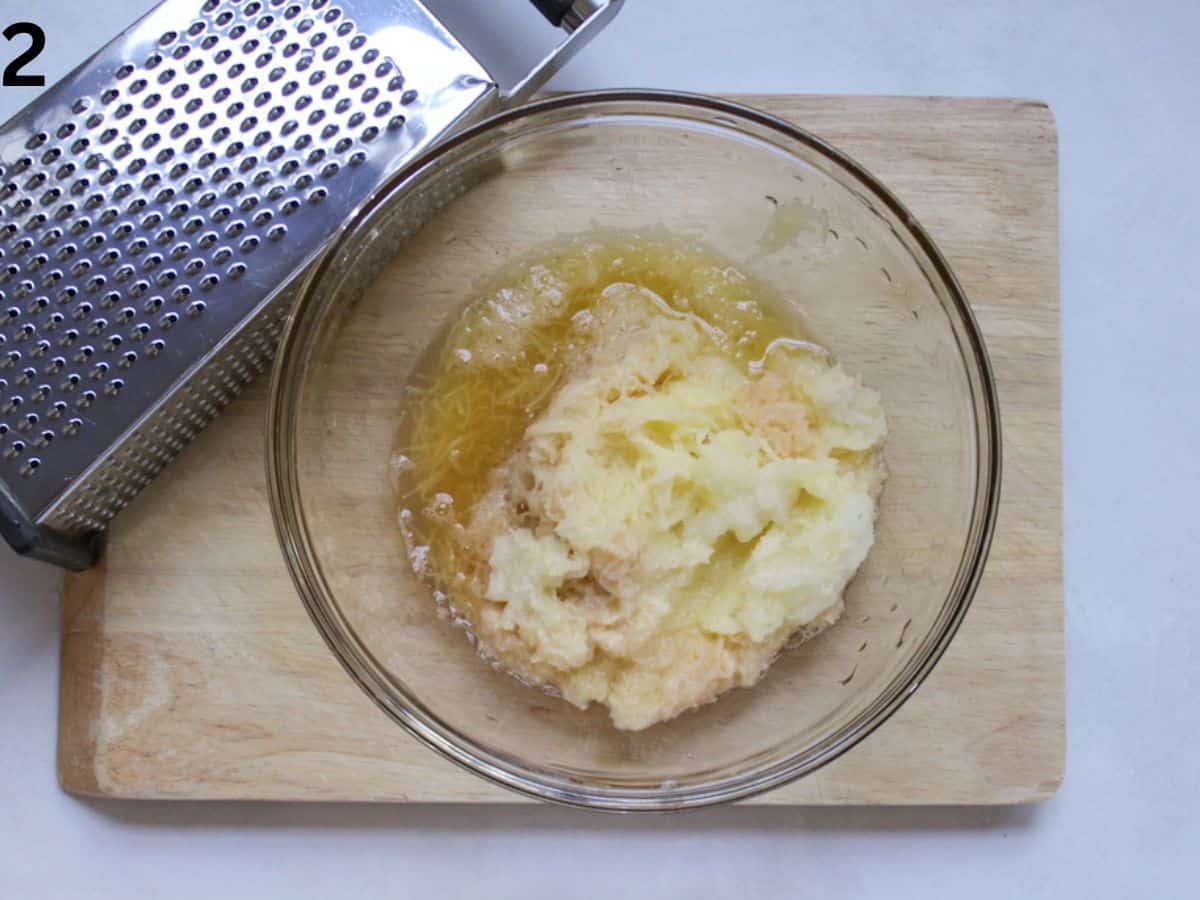 Grated potatoes in a glass bowl. There is a stainless steel box grater next to the bowl.