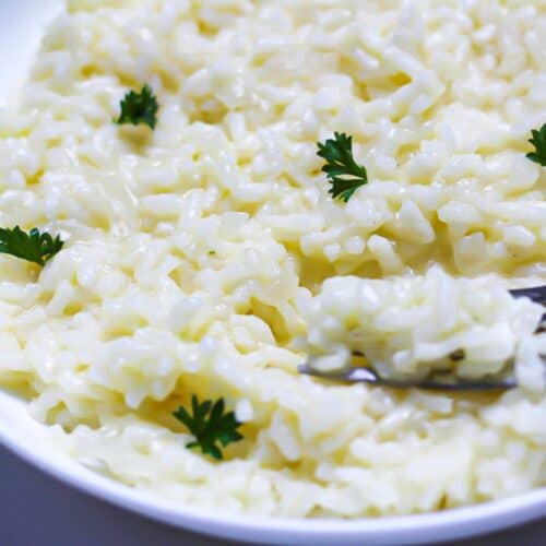 Creamy risotto in a plate with a fork scooping some. There is few fresh parsley leaves on top of the dish.