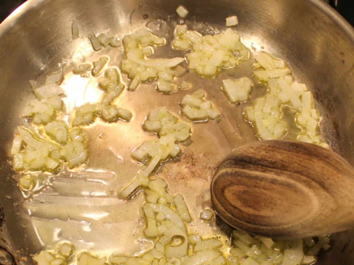 Finely diced onions are sautéed in a stainless steel pan. The wooden spoon is in the pan.