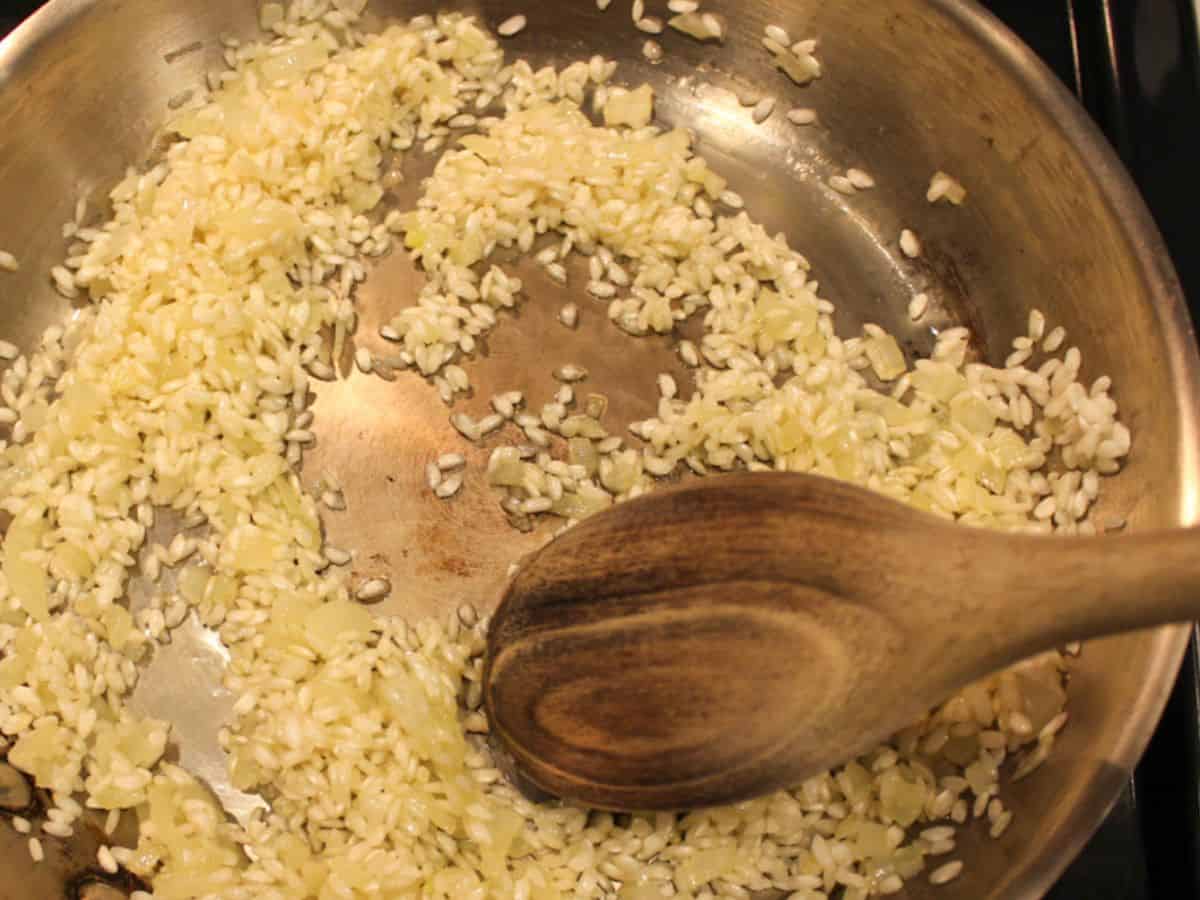 Arborio rice is toasted in a pan with onions with a wooden spoon in it for stirring.