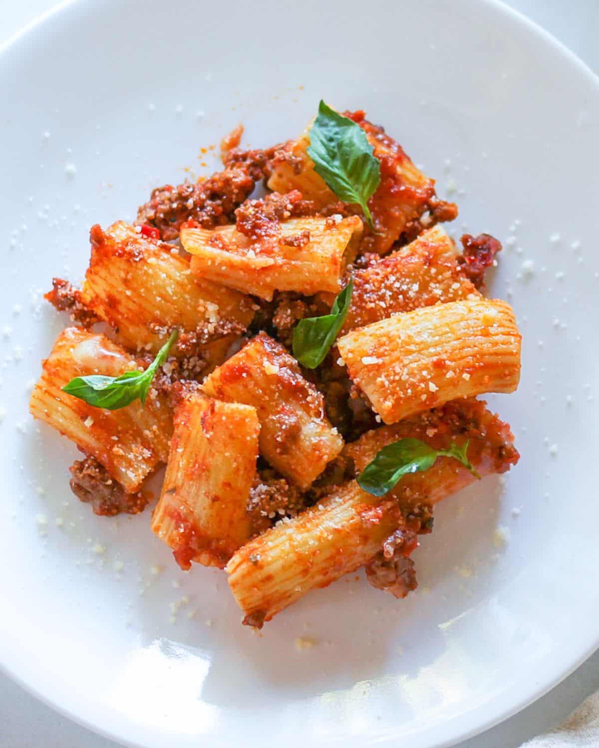 Baked Rigatoni pasta with tomato meat sauce on a white plate garnished with fresh basil leaves. 