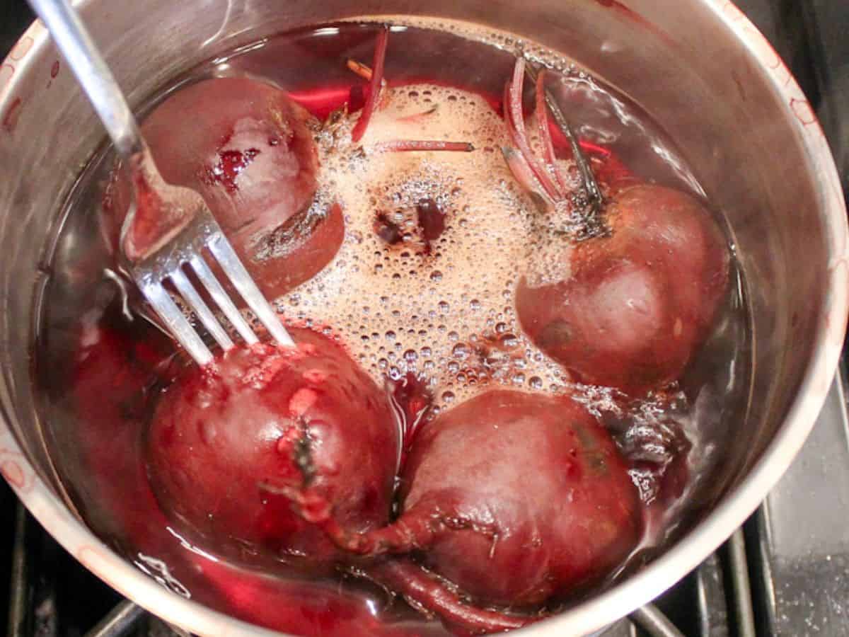 Four whole beets in pot with boiling water and a fork piercing through one beetroot.