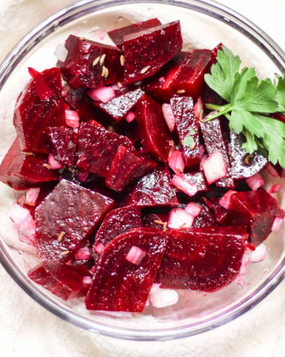 Rote Beete salat made with cooked red sliced beets, finenly diced onion and garnished with a green parsley leaf in a glass bowl.