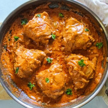 Reddish-orange chicken paprikash thighs in a large skillet garnished with green parsley on top.