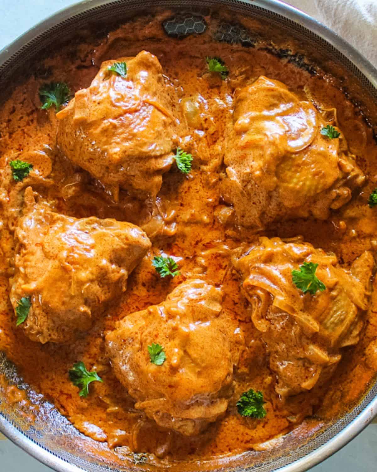 Reddish-orange chicken paprikash thighs in a large skillet garnished with green parsley on top.