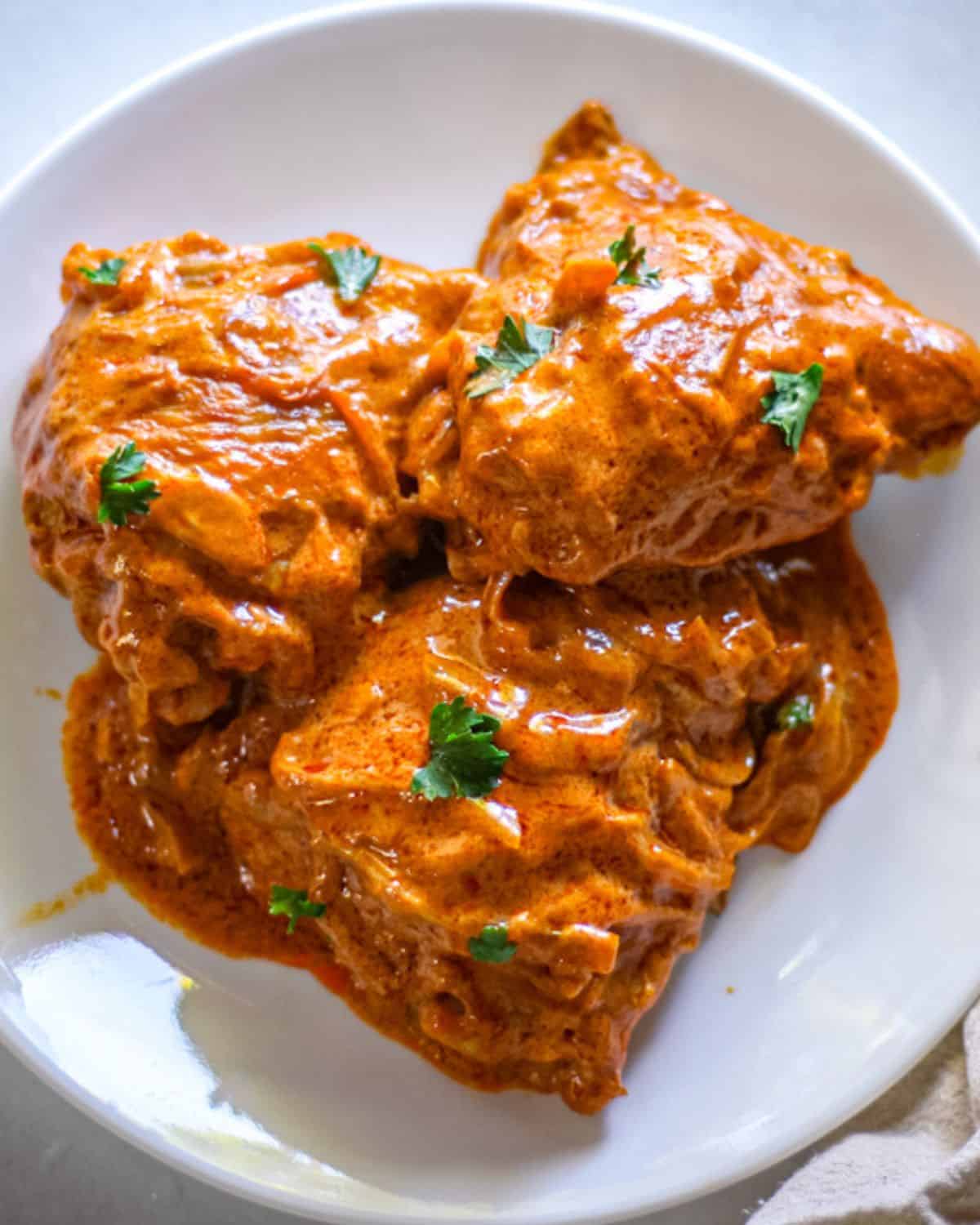 Chicken paprika thighs with orange sauce and some green parsley pieces on a white dinner plate.