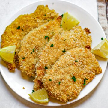 Four panko-crusted chicken breasts on a white plate with lemon wedges on the side and small pieces of chopped parsley on top.