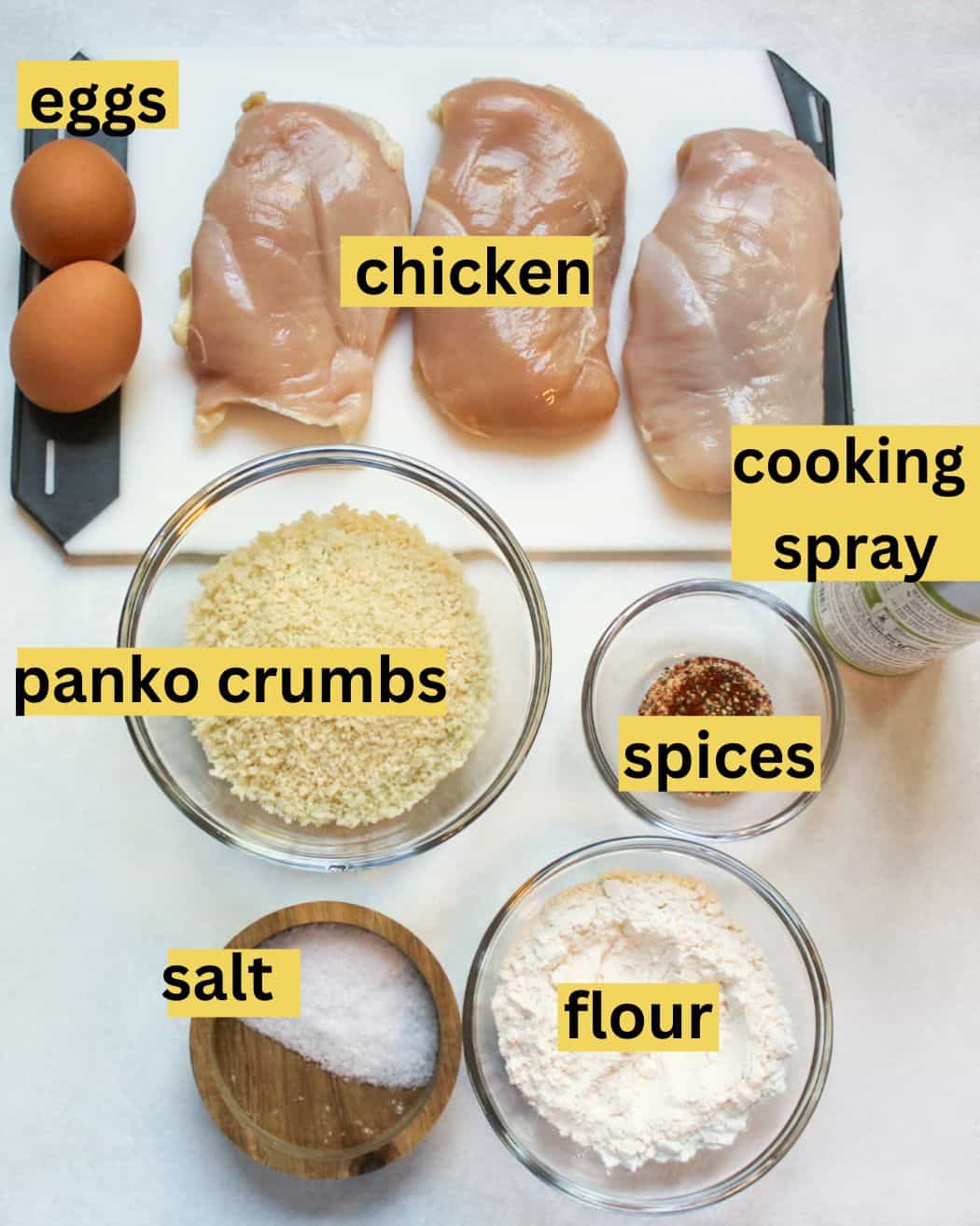 Recipe ingredients arranged on a white background labeled from top to bottom as eggs, chicken, cooking spray, panko crumbs, spices, salt, and flour.