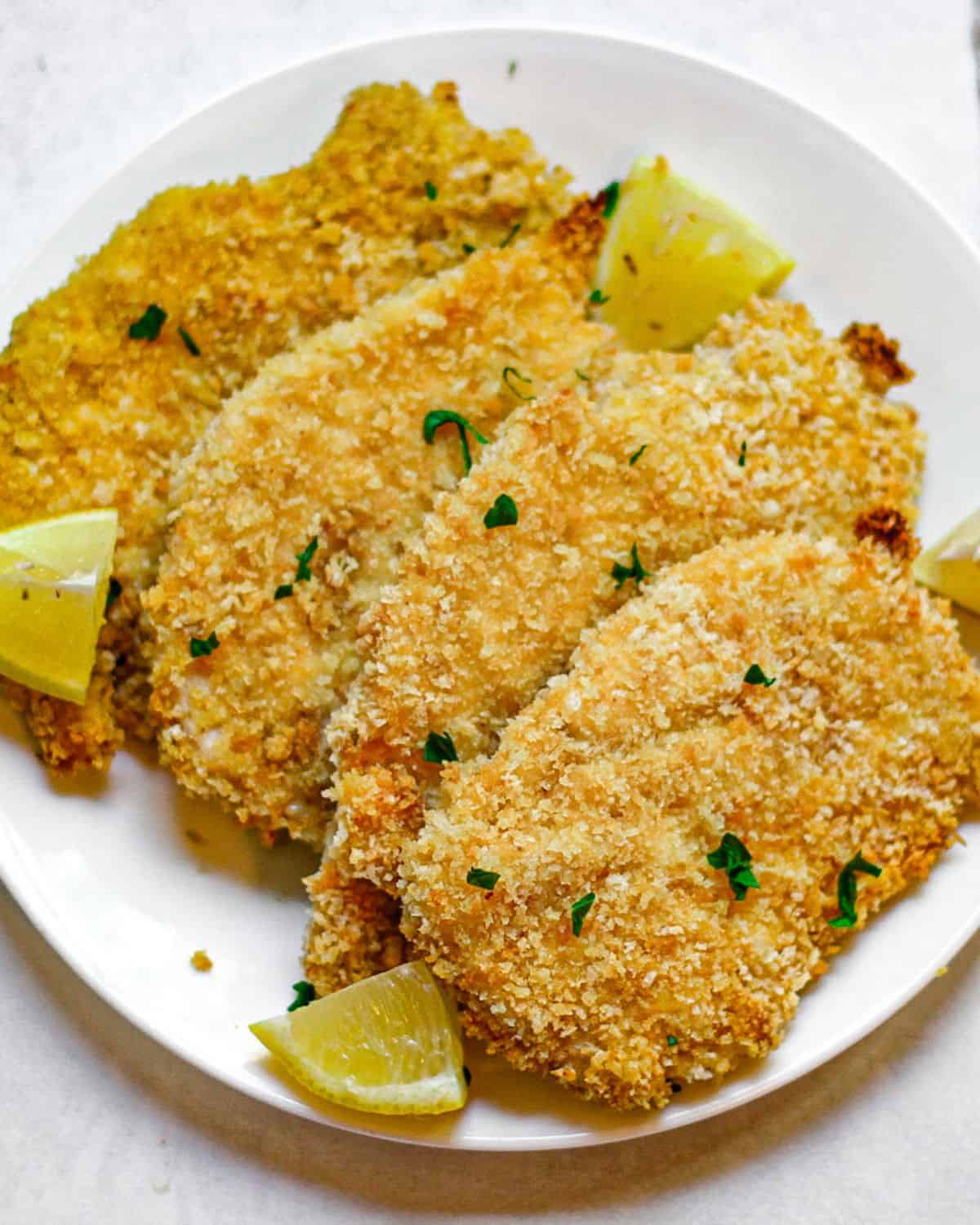 Four panko-crusted chicken breasts on a white plate with lemon wedges on the side and small pieces of chopped parsley on top. 