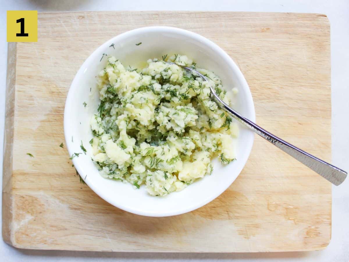 Mashed potatoes mixed with fresh herbs in a white bowl with a spoon in it. 