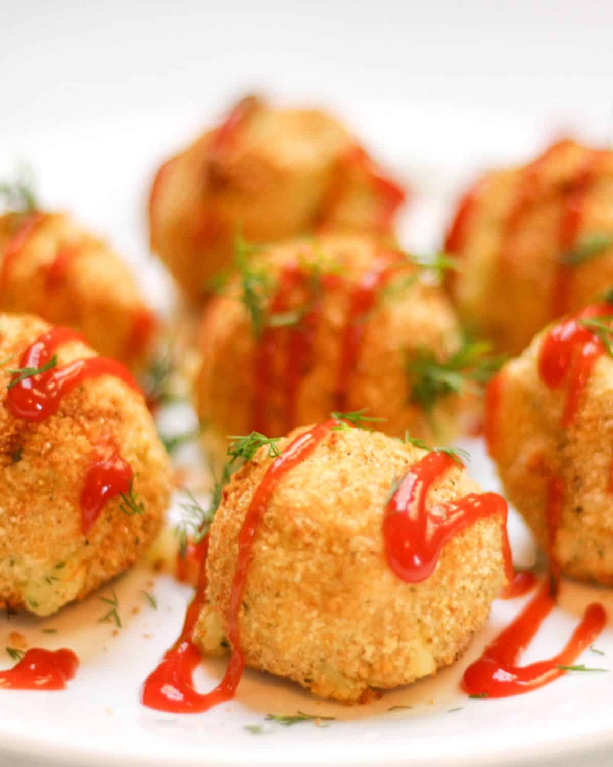 Mashed potato croquettes shaped as balls on a white plate drizzled with ketchup on top and sprinkled with fresh dill.