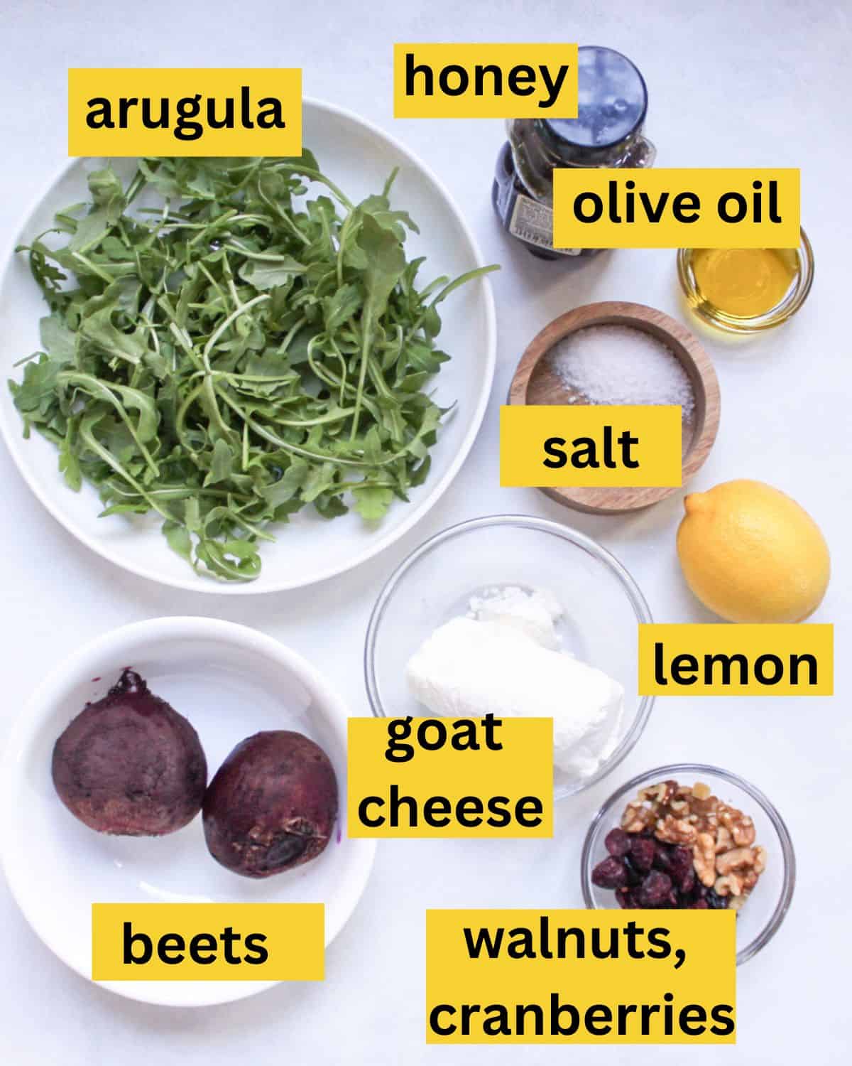 Salad recipe ingredients on a white background labeled as arugula, olive oil, honey, salt, goat cheese, lemon, beets, cranberries and walnuts.