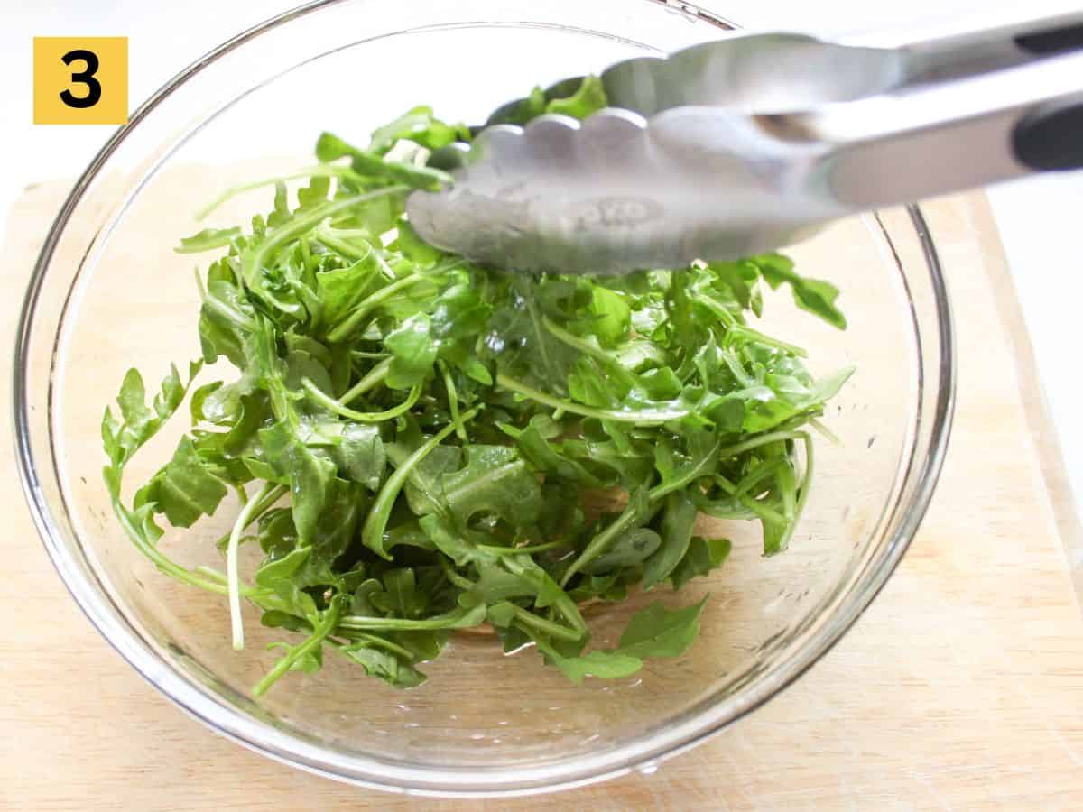 Arugula leaves in a large glass bowl being tossed in the dressing with tongs.