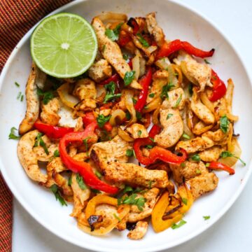 Cooked chicken fajitas with onions and peppers in a white plate garnished with chopped green cilantro and half of the lime on the side.