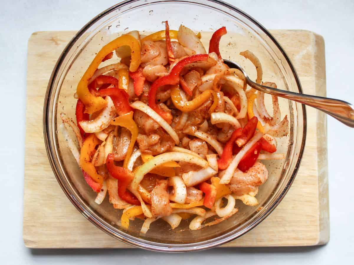 Raw strips of chicken, sliced peppers, red and yellow bell peppers in a large glass bowl.