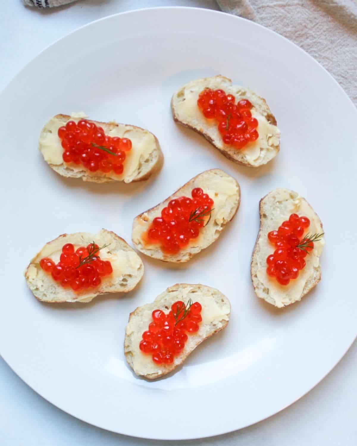Six buttered baguette slices topped with red caviar on a white plate.