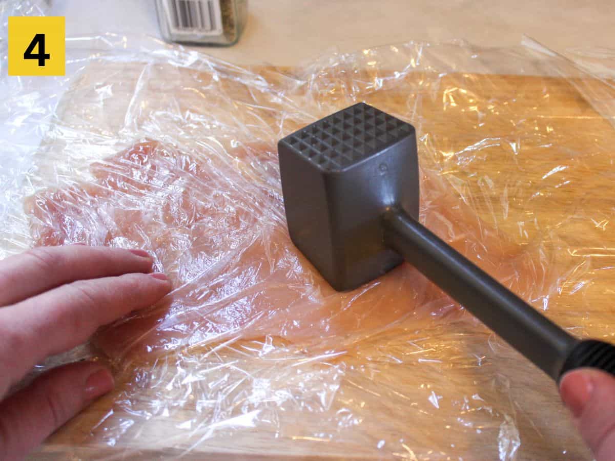 Pounding the chicken covered with a flat side of meat tenderizer. The chicken is covered with plastic wrap.