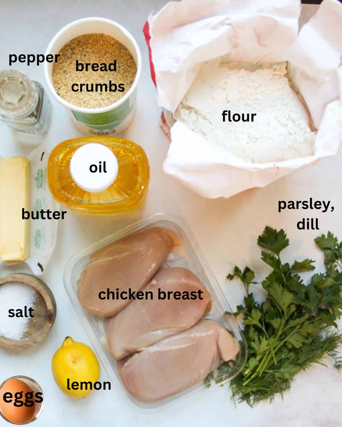 Recipe ingredients on a white surface labeled as salt, butter, breadcrumbs, oil, chicken breast, flour parsley, eggs, lemon, dill.