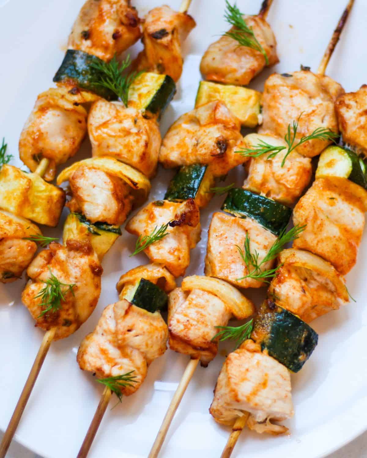 Baked chicken and vegges skewers on a white place garnished with fresh herbs on top. 