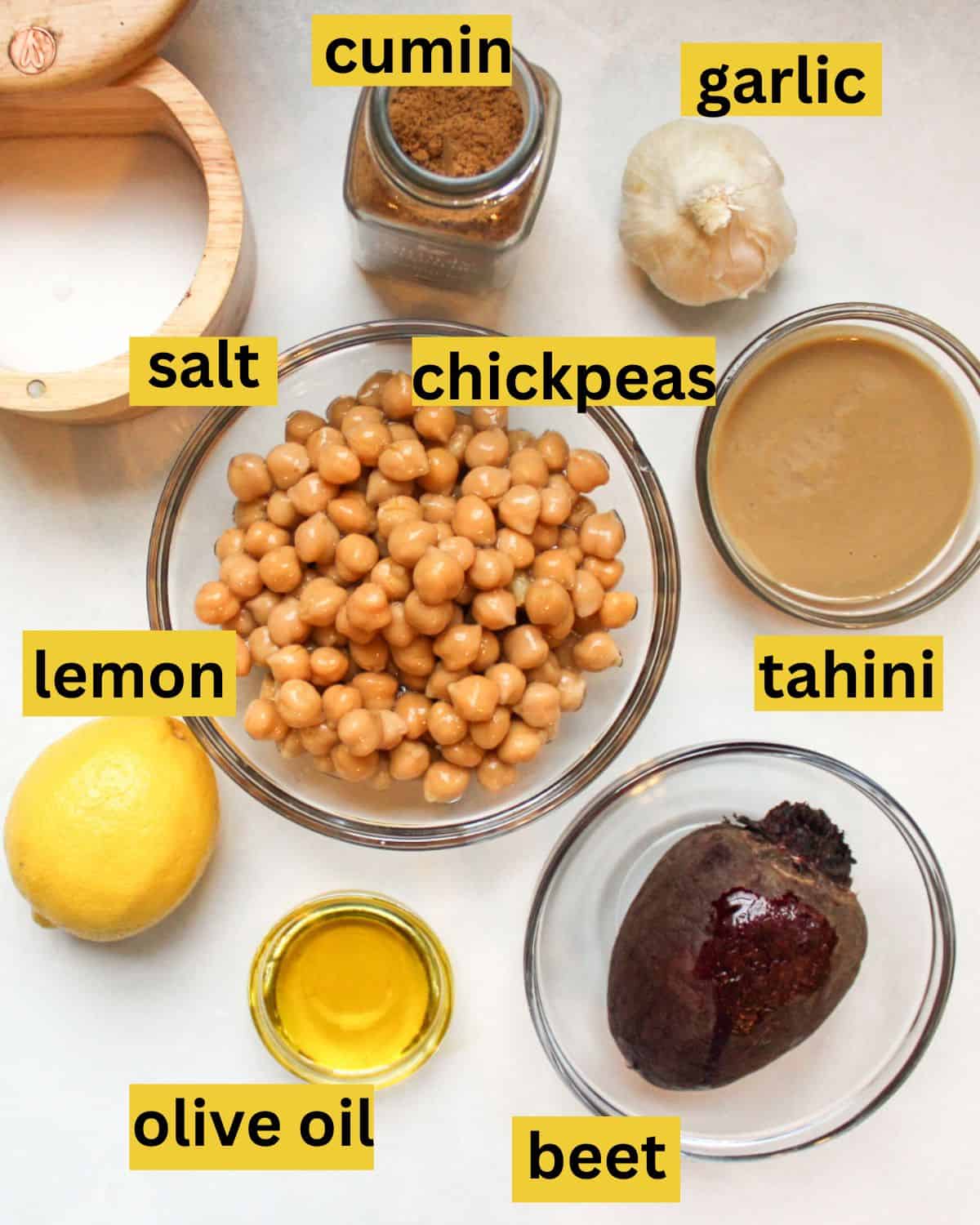 All the recipe ingredients neatly arranged on a white background, each labeled salt, lemon, cumin. chickpeas, olive oil, garlic, tahini, beet.