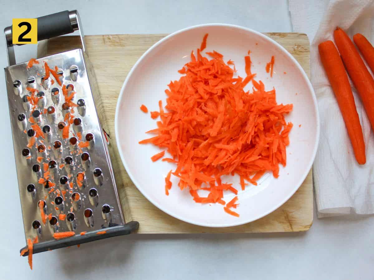A shallow dish filled with grated carrots , accompanied by a box grater at its side.