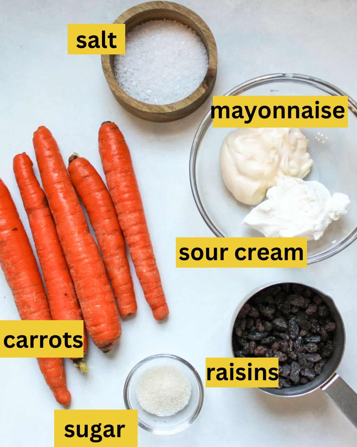 All the recipe ingredients neatly arranged on a white background, each labeled as carrots, salt, mayonnaise, sour cream, sugar, raisins.