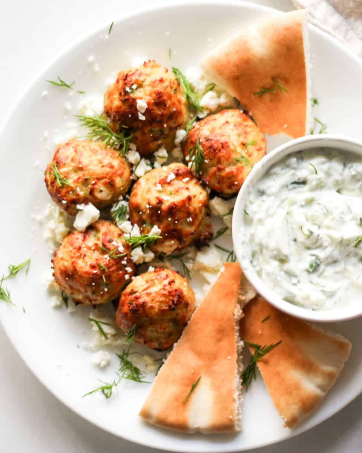 Greek chicken meatballs sprinkled with fresh dill on a white plate with pita bread and tzatziki sauce on the side. 