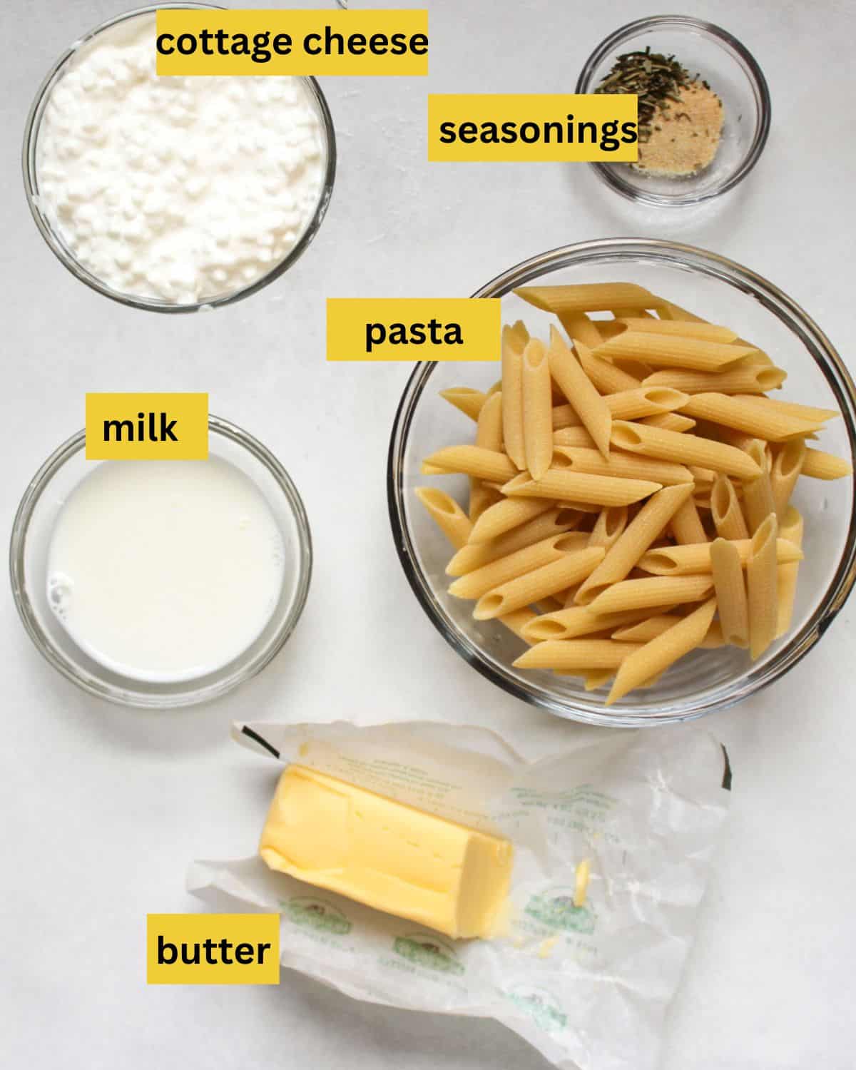 All the recipe ingredients neatly arranged on a white background, each labeled as cottage cheese, seasonings, milk, pasta. and butter.