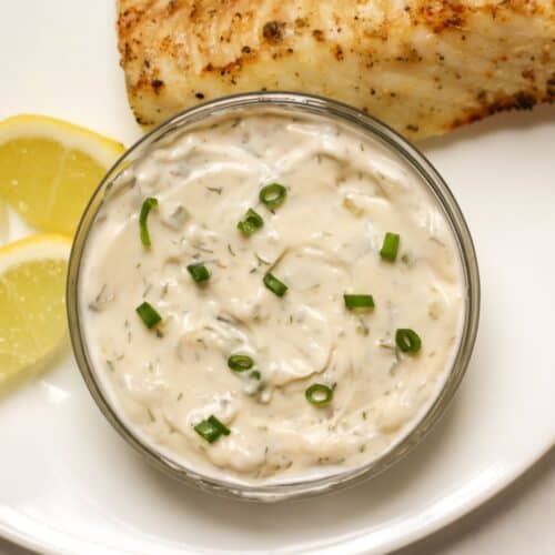 Homemade tartar sauce in a glass bowl garnished with chopped green onions, served with fried fish and lemon wedges on a plate.