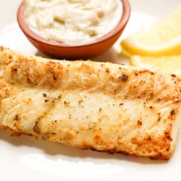 Prepared cod fillet on a white dish accompanied by tartar sauce and a lemon wedge on the side.