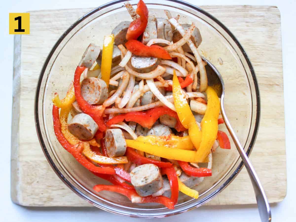 Thinly sliced onions, bell peppers and sausage in a large glass bowl with a spoon in it.