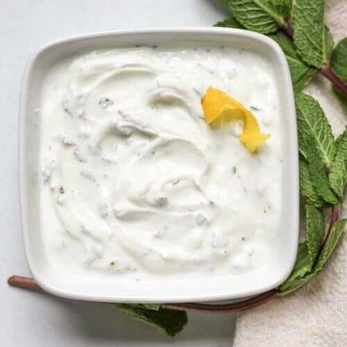 Square white bowl filled with mint Greek yogurt sauce, garnished with a lemon peel and fresh mint leaves placed next to the bowl.