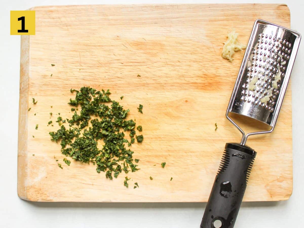 On a wooden cutting board, finely chopped mint is next to a small cheese grater with freshly grated garlic next to it.