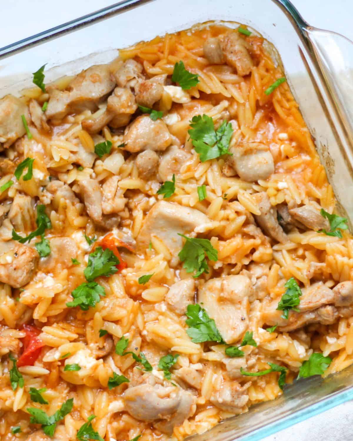 Baked chicken and orzo casserole garnished with chopped green herbs on top in a glass square dish.