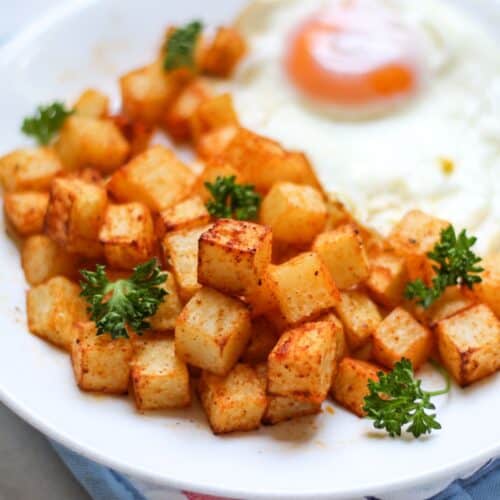 Air-fried potato cubes garnished with fresh curly parsley served with a sunny-side-up egg on a white plate.