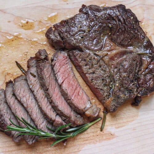 Sliced cooked steak with slightly pink center on a cutting board, garnished with with a fresh sprig of rosemary.