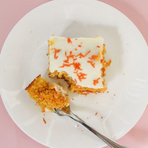 Overhead view of a square slice of carrot semolina cake with white glaze and shredded fresh carrots on top, served on a white plate with a bite-size piece on a fork showing the soft golden crumb inside.