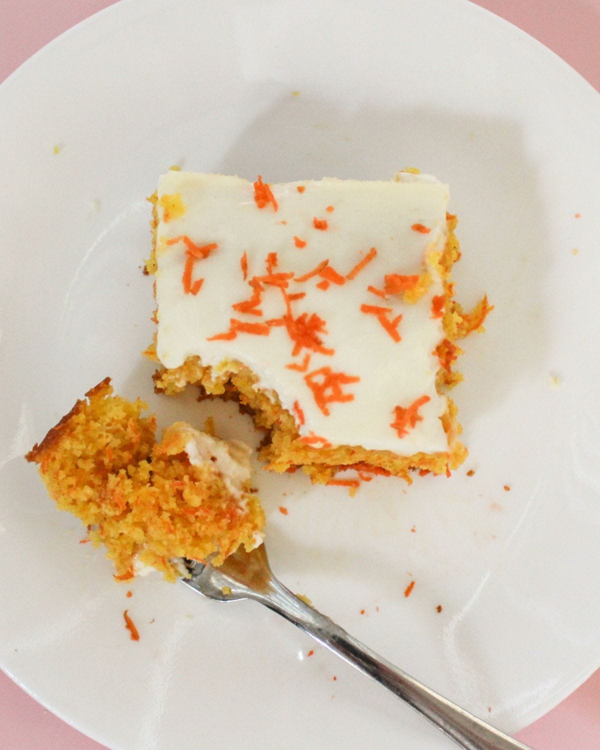 Overhead view of a square slice of carrot semolina cake with white glaze and shredded fresh carrots on top, served on a white plate with a bite-size piece on a fork showing the soft golden crumb inside.