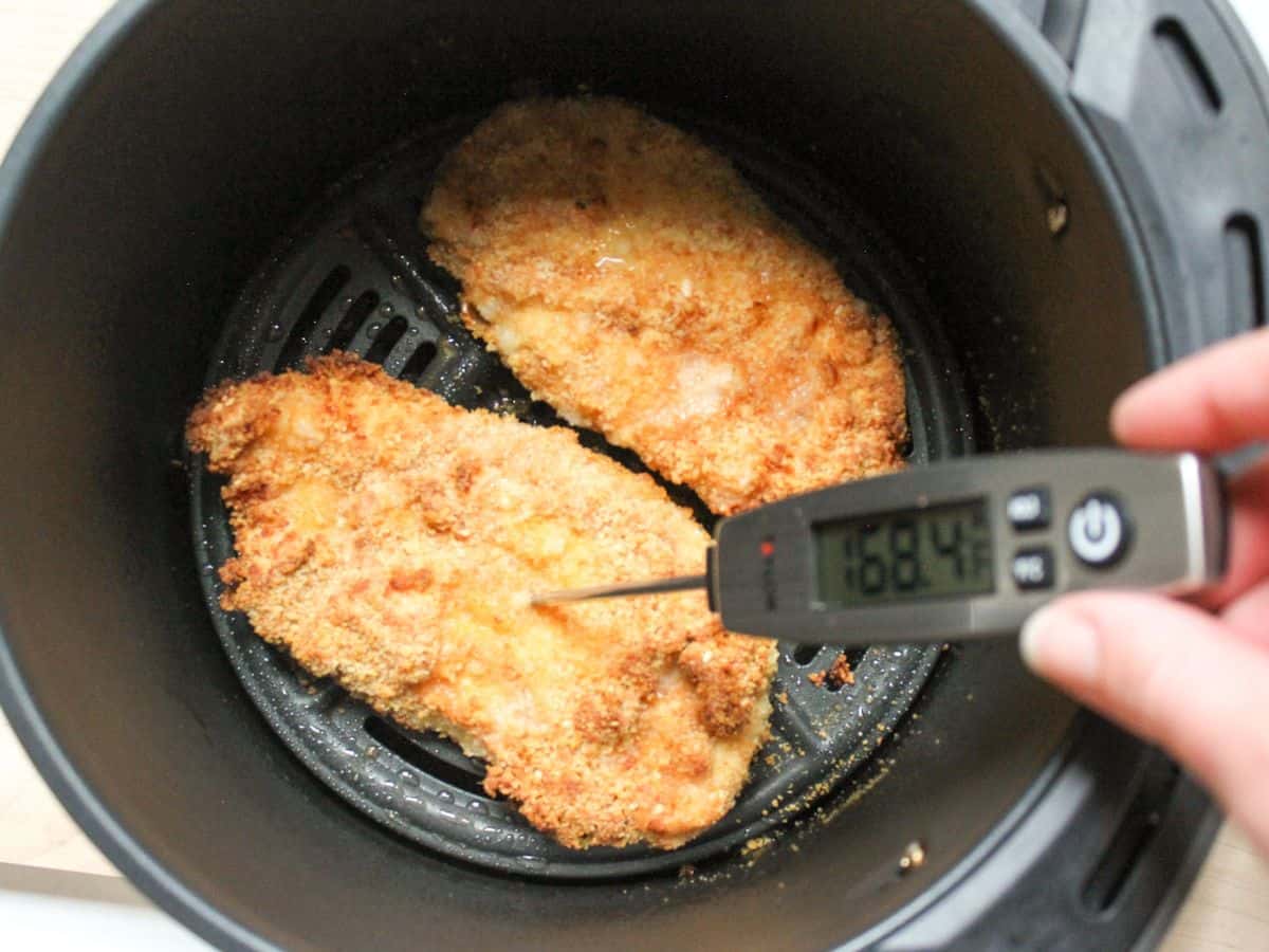Two cooked chicken schnitzels in air fryer basket with meat thermometer in one of them checking internal temperature.