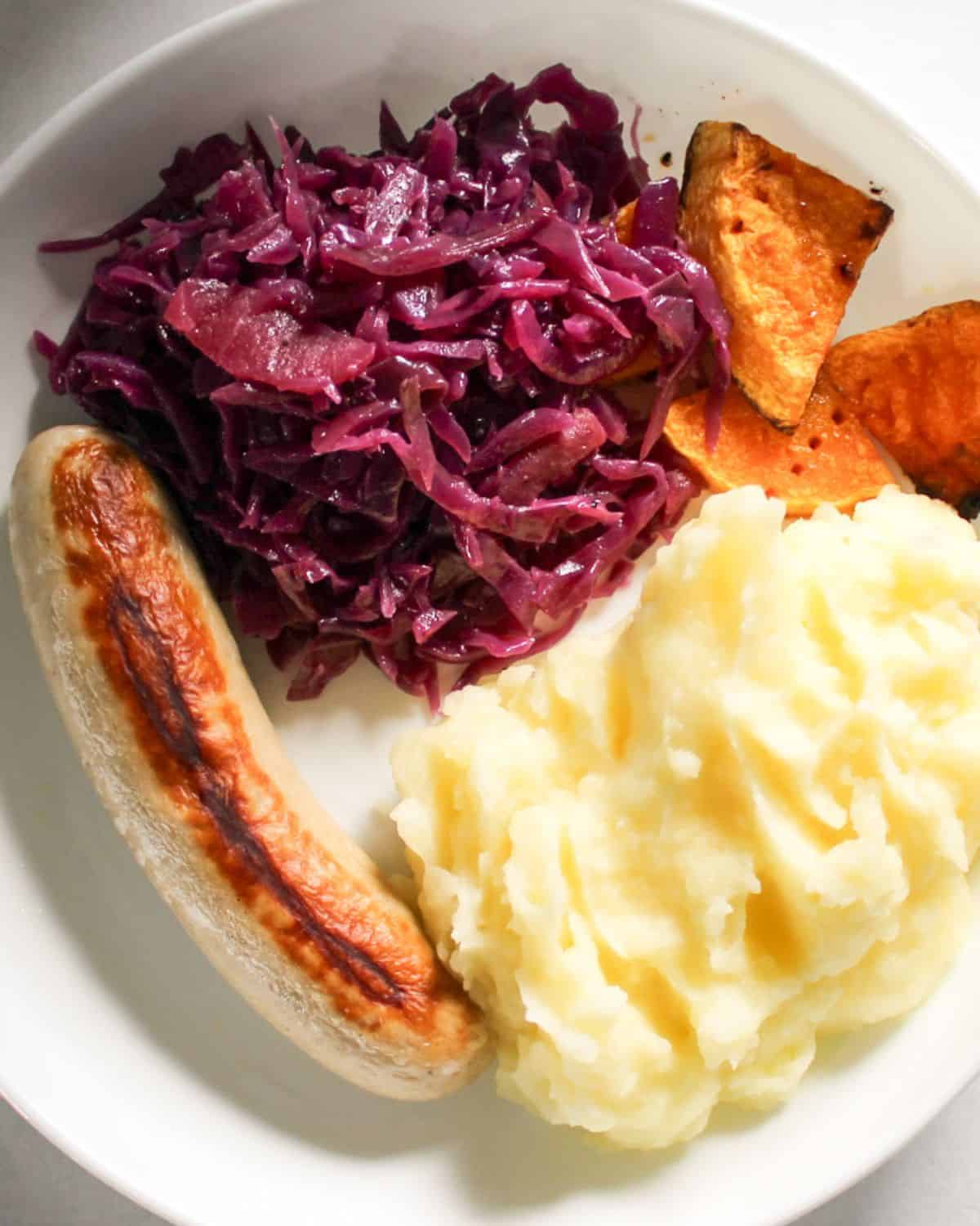Red braised cabbage served with mashed potatoes, bratwurst on a white plate.