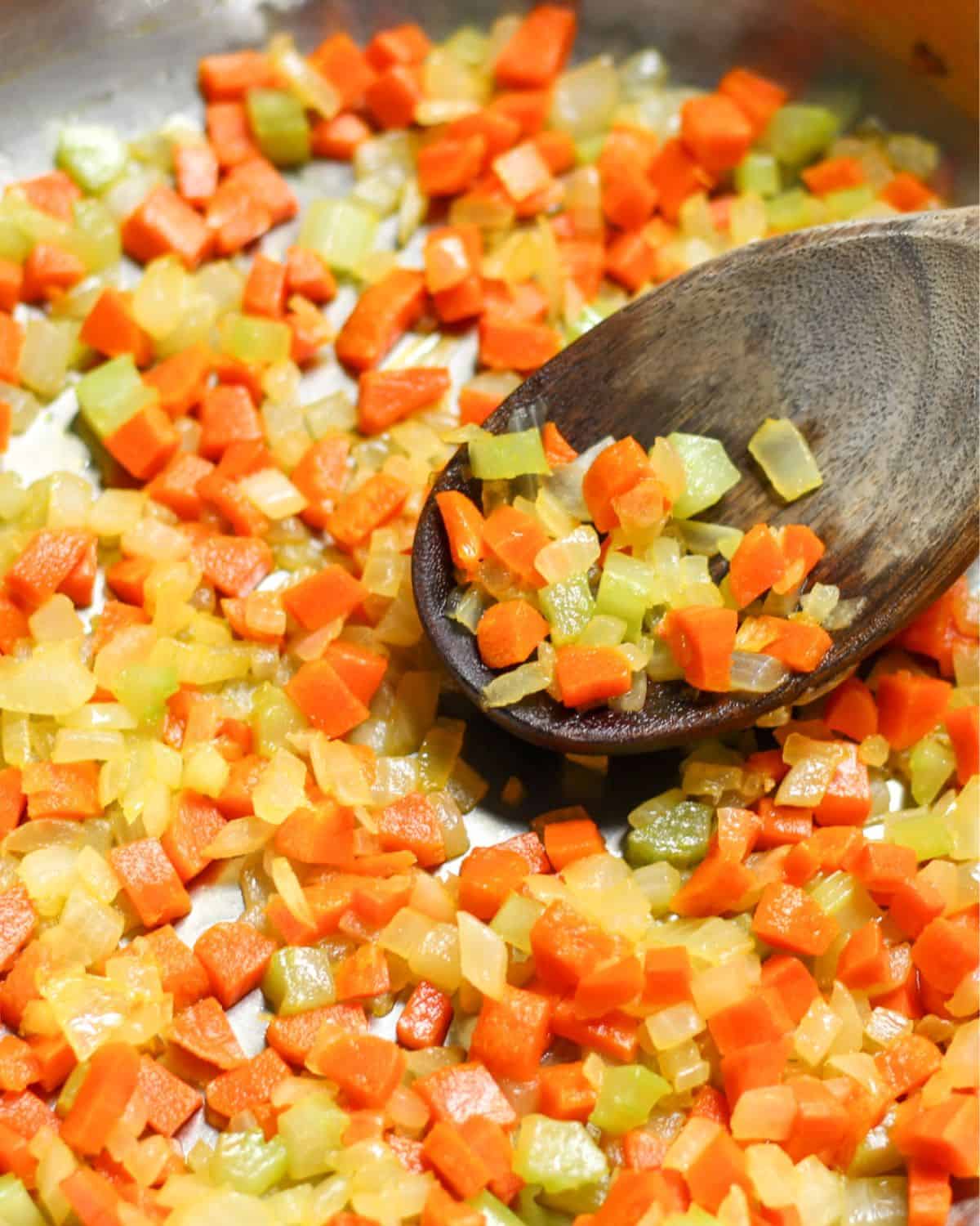 Sauteed mirepoix with diced onions, carrots, celery in a skillet being scooped with wooden spoon.