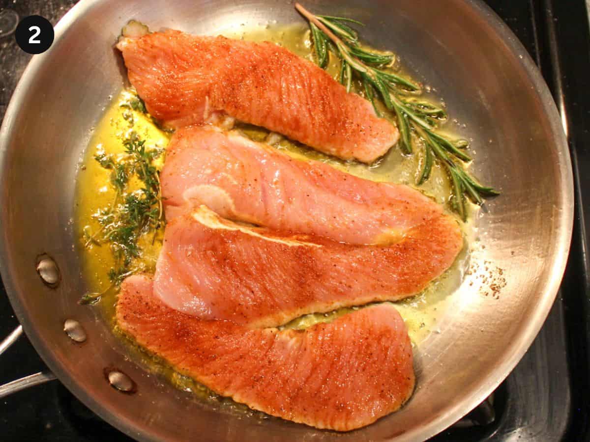 Searing turkey steaks in stainless steel frying pan with added fresh rosemary and thyme.